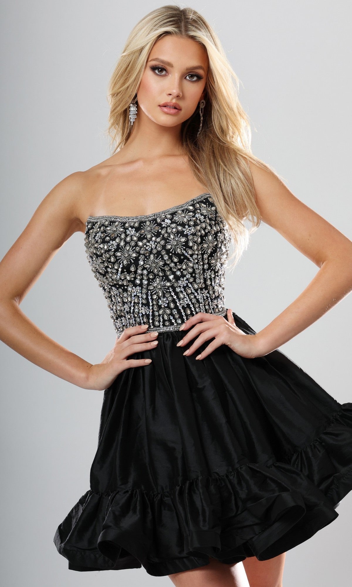 A woman poses in the Jovani 52439 short homecoming dress in black/silver, featuring a strapless, beaded and embellished bodice with a ruffled skirt, set against a plain background.